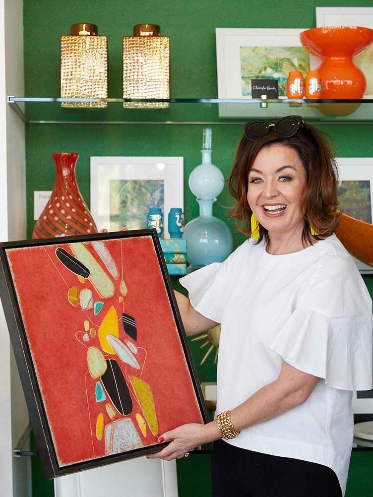 Anna Brockway poses with orange abstract painting in front of glass shelves.