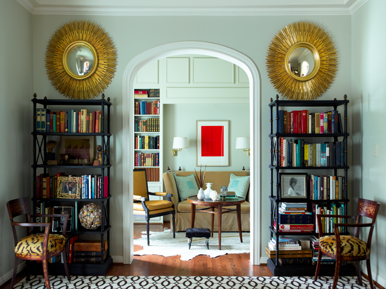 Twin black bookshelves, gold-burst round mirrors, and wood accent chair with tiger cushions on either side of an archway