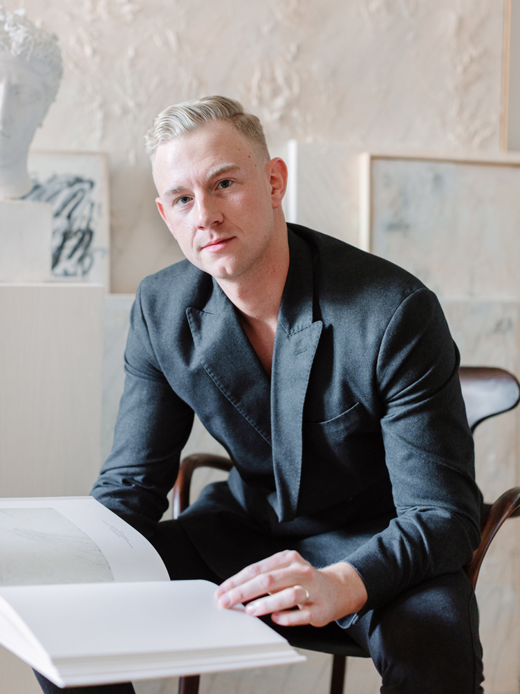 Artist Josh Yöung sits on a mid-century wooden chair reading an art book.