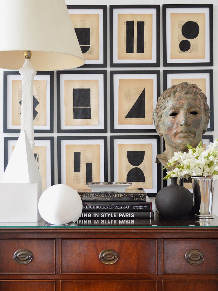 A wall of contemporary black, white, and beige art is framed above a large wooden chest with sculptures and books.