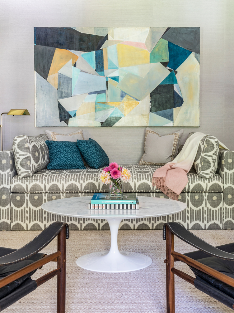 Abstract art hangs above a gray patterned couch with many pillows and a tulip coffee table.