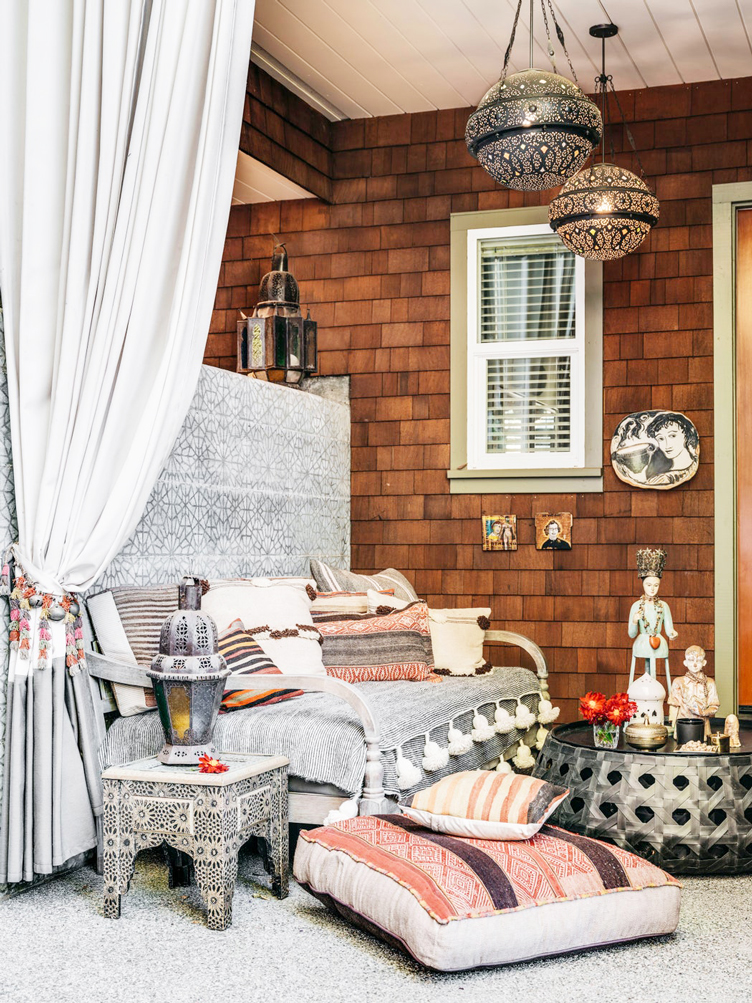 Maximalist outdoor patio combines brown shingling with gray textiles, floor pillows, and woven coffee table.