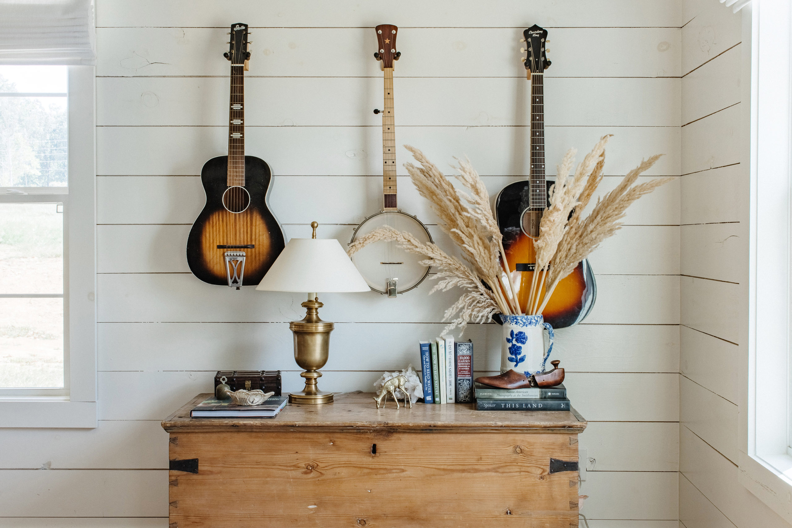 Wall-mounted string instruments hang above raw wood chest and vase of wheat.