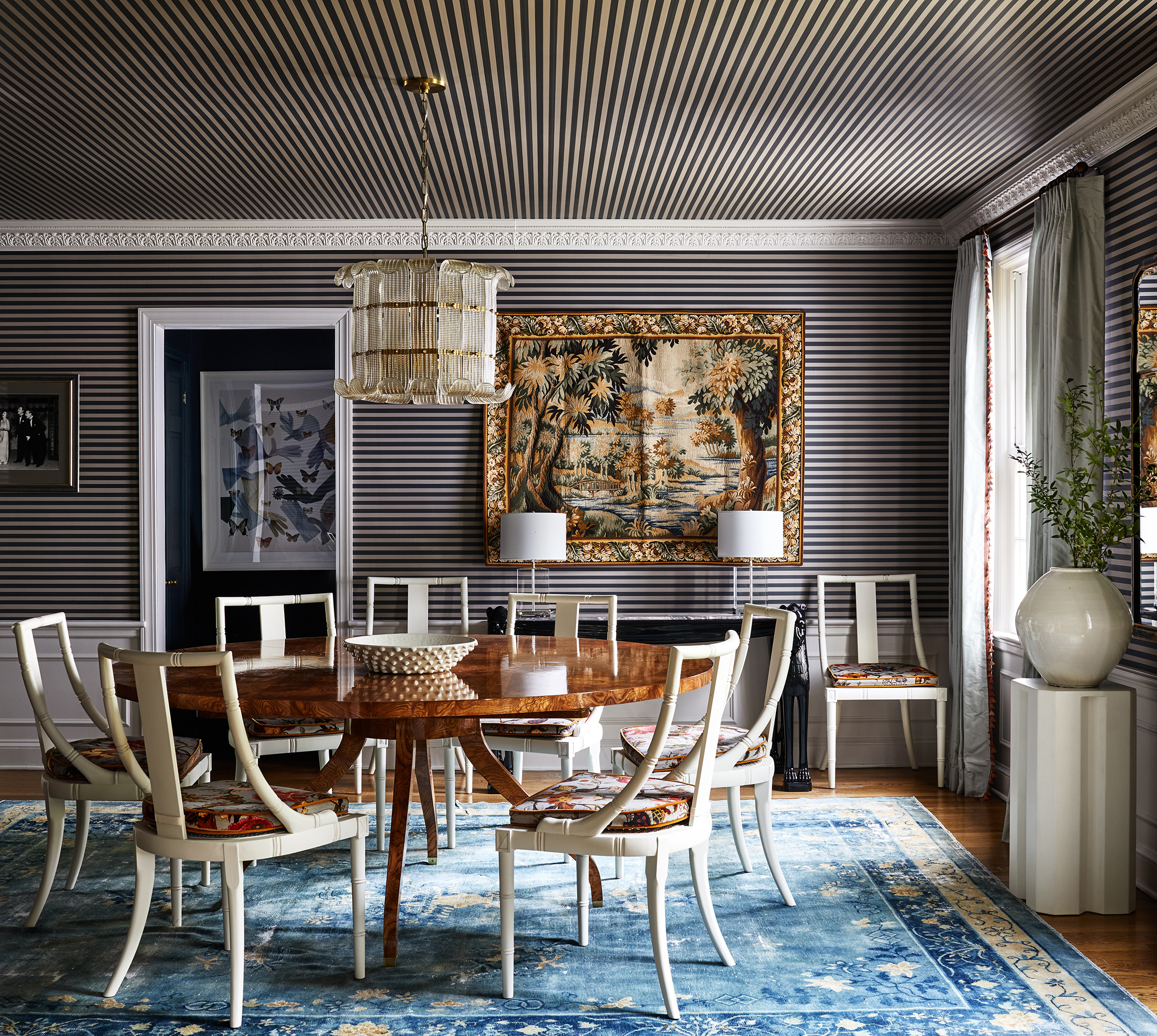 Eclectic dining room with round, burl wood table, striped wallpaper, and hanging tapestry.