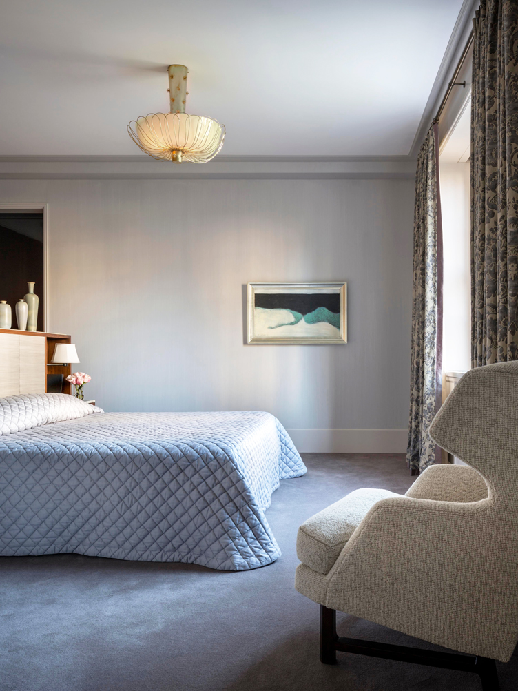 Bedroom with blue quilted blanket and cream wool armchair is paired with light blue walls.