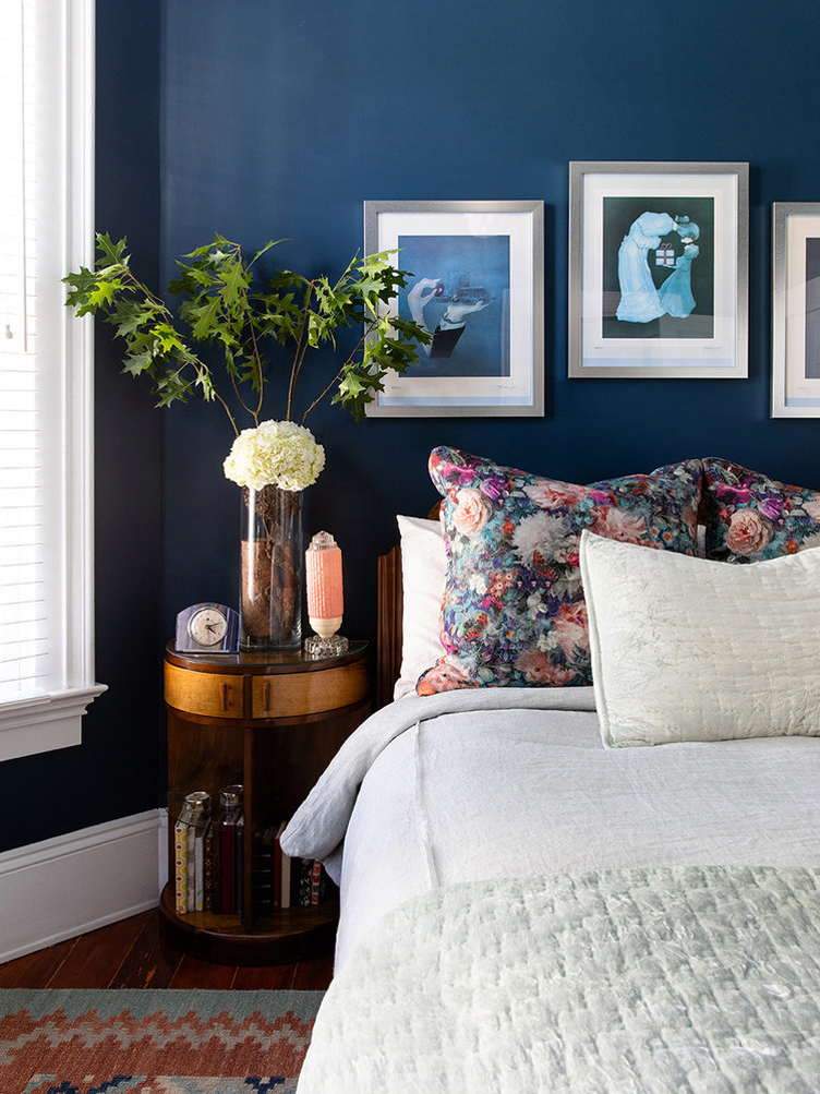 Dark blue painted walls compliment the blue wall art, floral throw pillows, and dark wood side table.