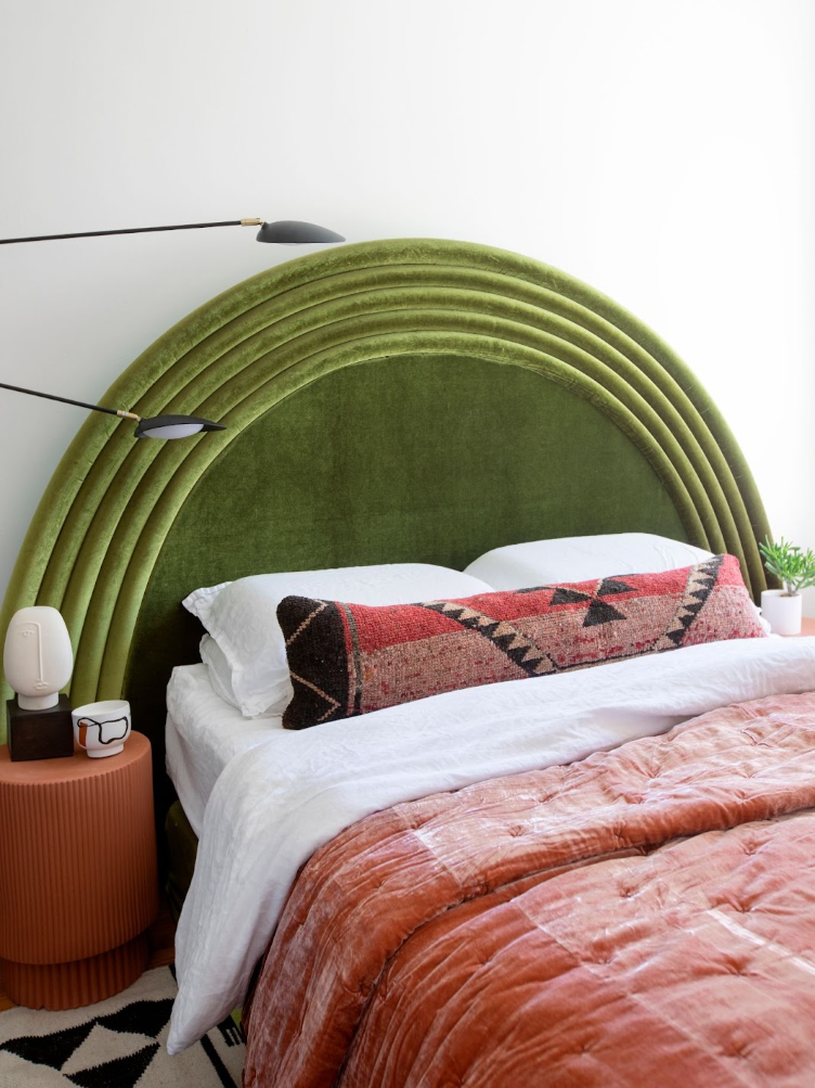 Bedroom with arched green, velvet headboard, woven throw pillow, and orange drum side table