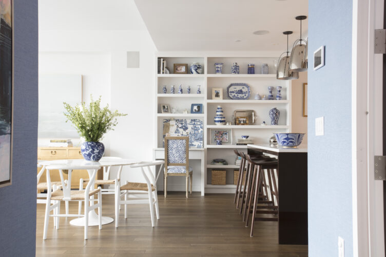 Dining area with white Scandinavian dining chairs and white tulip table beside white shelves with blue and white ceramic pieces.