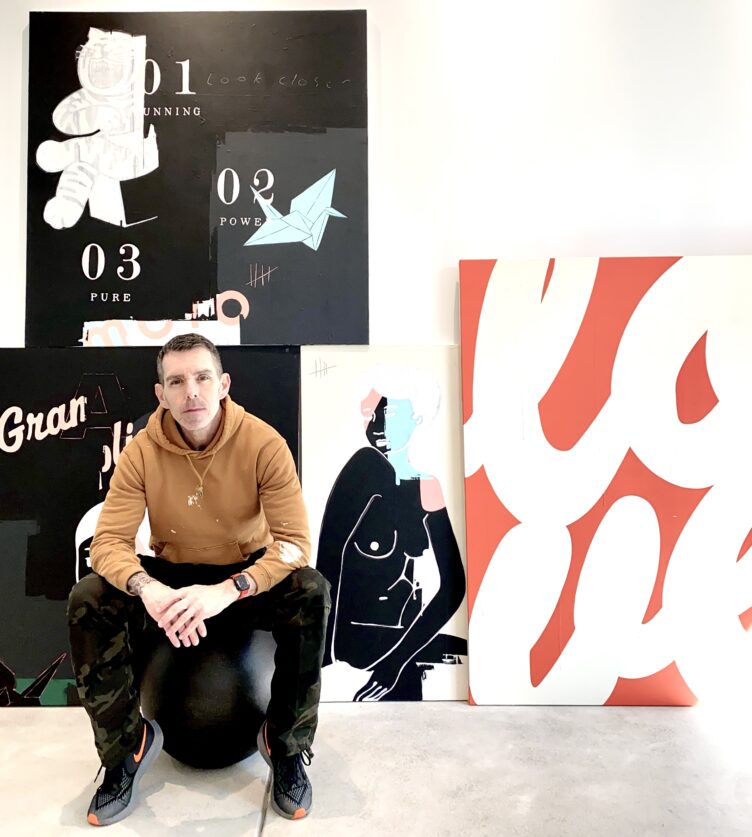 Ron Giusti poses on a black spherical ottoman in front of an array of his painting in black, white, pink, and orange.