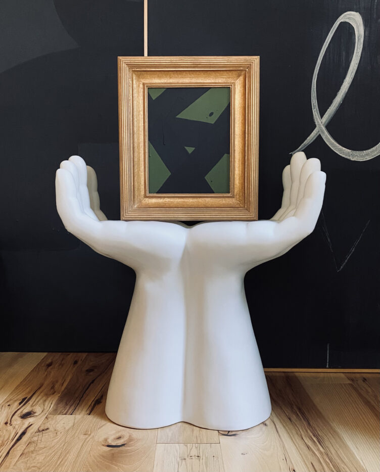 White hand sculpture holds gold framed abstract green and black painting.