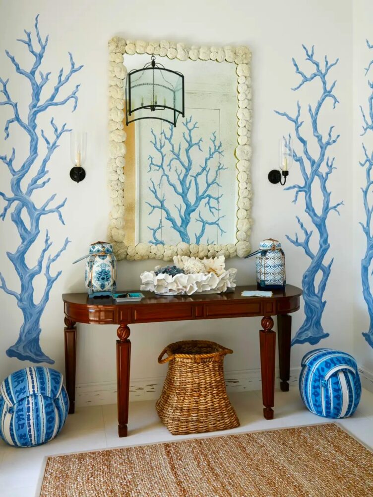 A wicker baskets sits below this console in a Palm Beach entry. Two poufs, ginger jars, and a seashell bordered mirror surround the wicker basket.