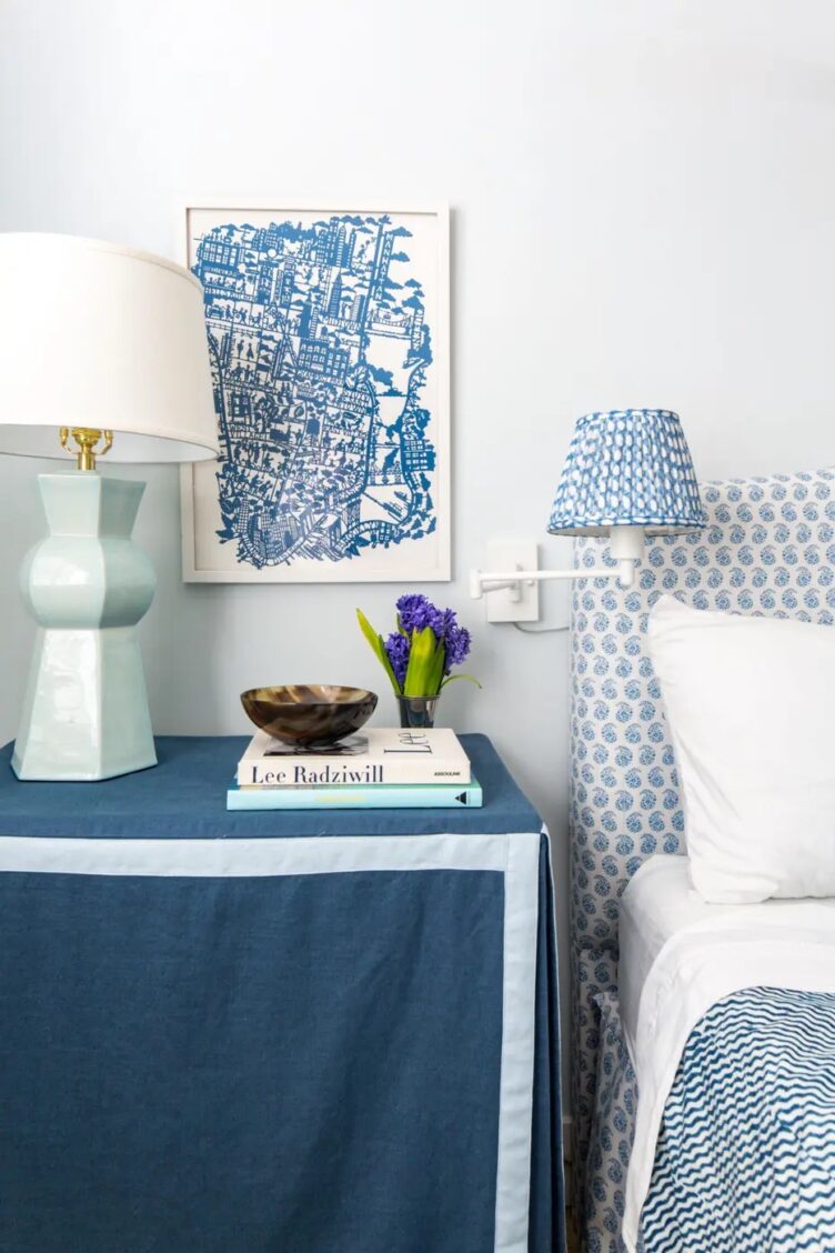 Nightstand dressed in navy blue piped skit, with aqua ceramic lamp, books, horn bowl, and hyacinths in a silver julep cup